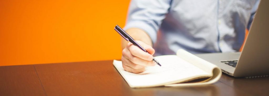 A man's hand is holding a pen and it is poised over a pad of paper