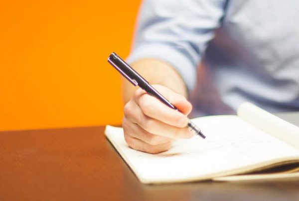 A man's hand is holding a pen and it is poised over a pad of paper