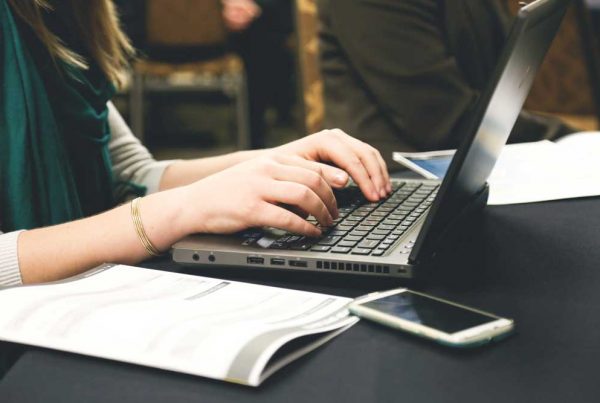 A woman is minute taking on a laptop, a phone and some paper are on the table