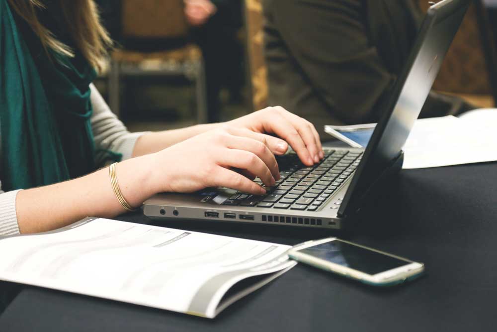 A woman is minute taking on a laptop, a phone and some paper are on the table