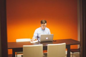 A man sits at a table with a laptop in front of him, taking notes on a paper note pad